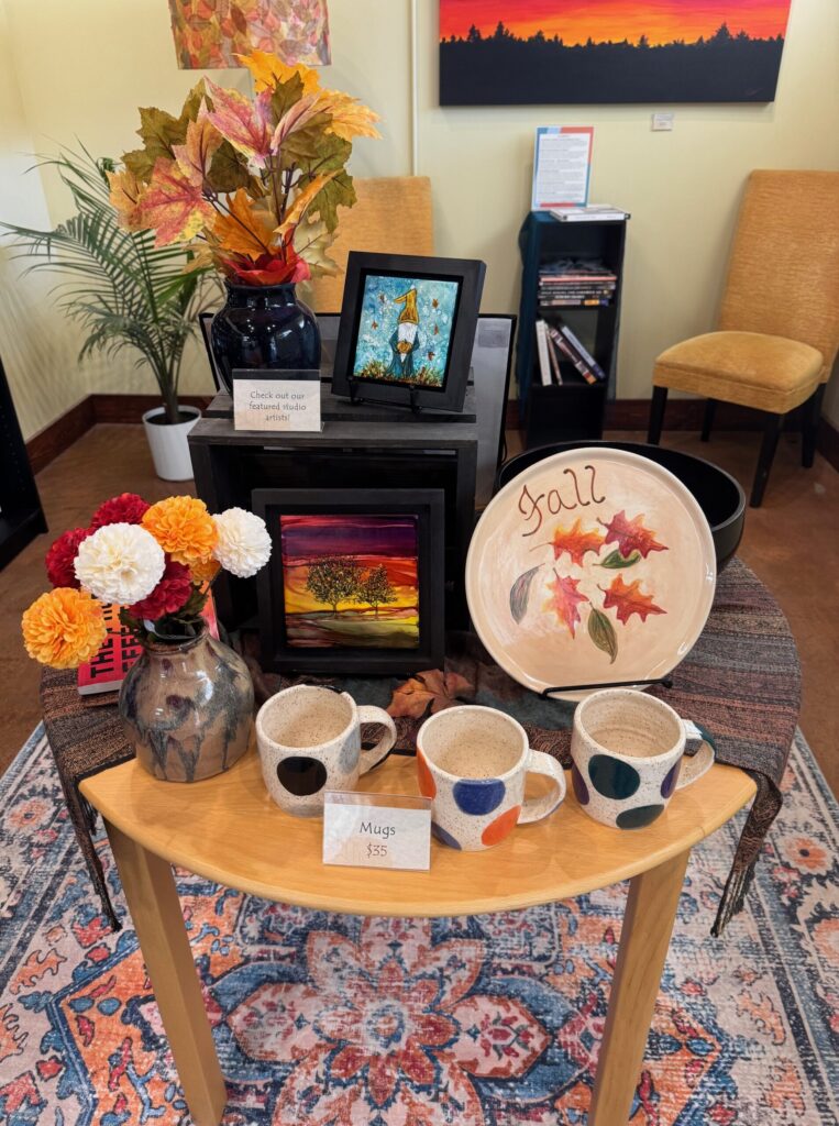 Display of handmade pottery mugs, fall-themed plate, framed artwork, and floral arrangements in the House of Clay gallery in Overland Park.